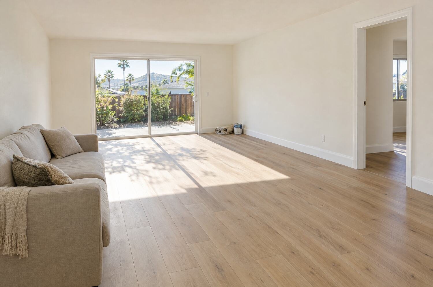 Finished living room with new light oak plank flooring, fresh warm white walls and white baseboards, a rolled drop cloth and paint can in the corner